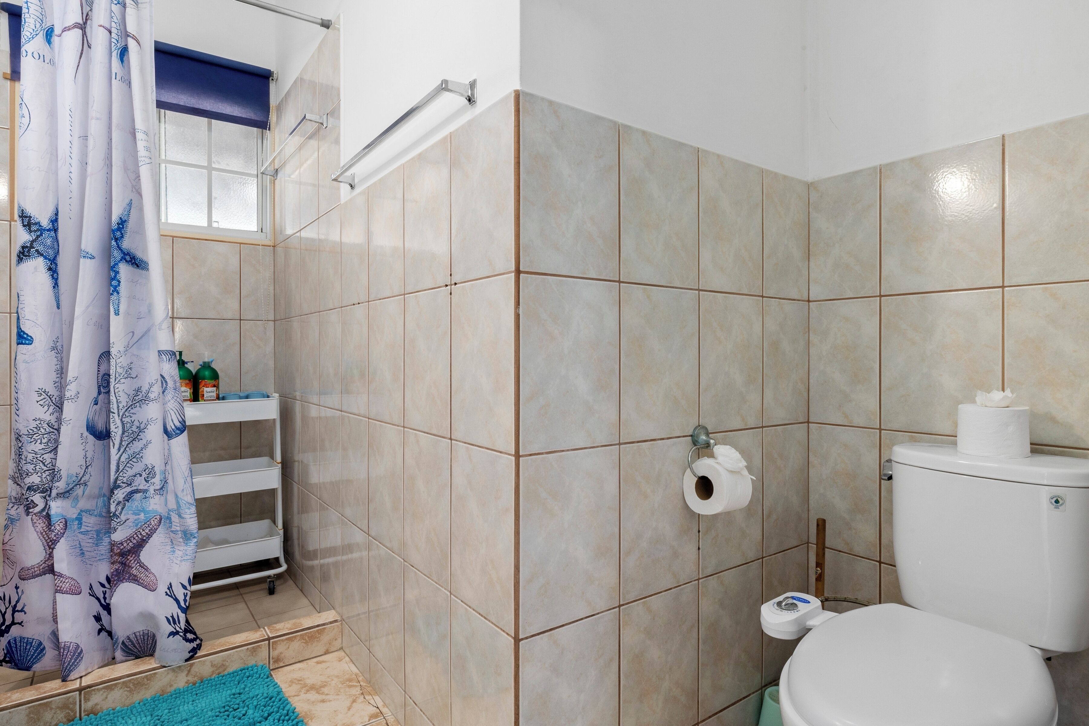 A bathroom with beige tiles, a white toilet, a blue shower curtain with starfish and coral patterns, and a small shelf with toiletries.