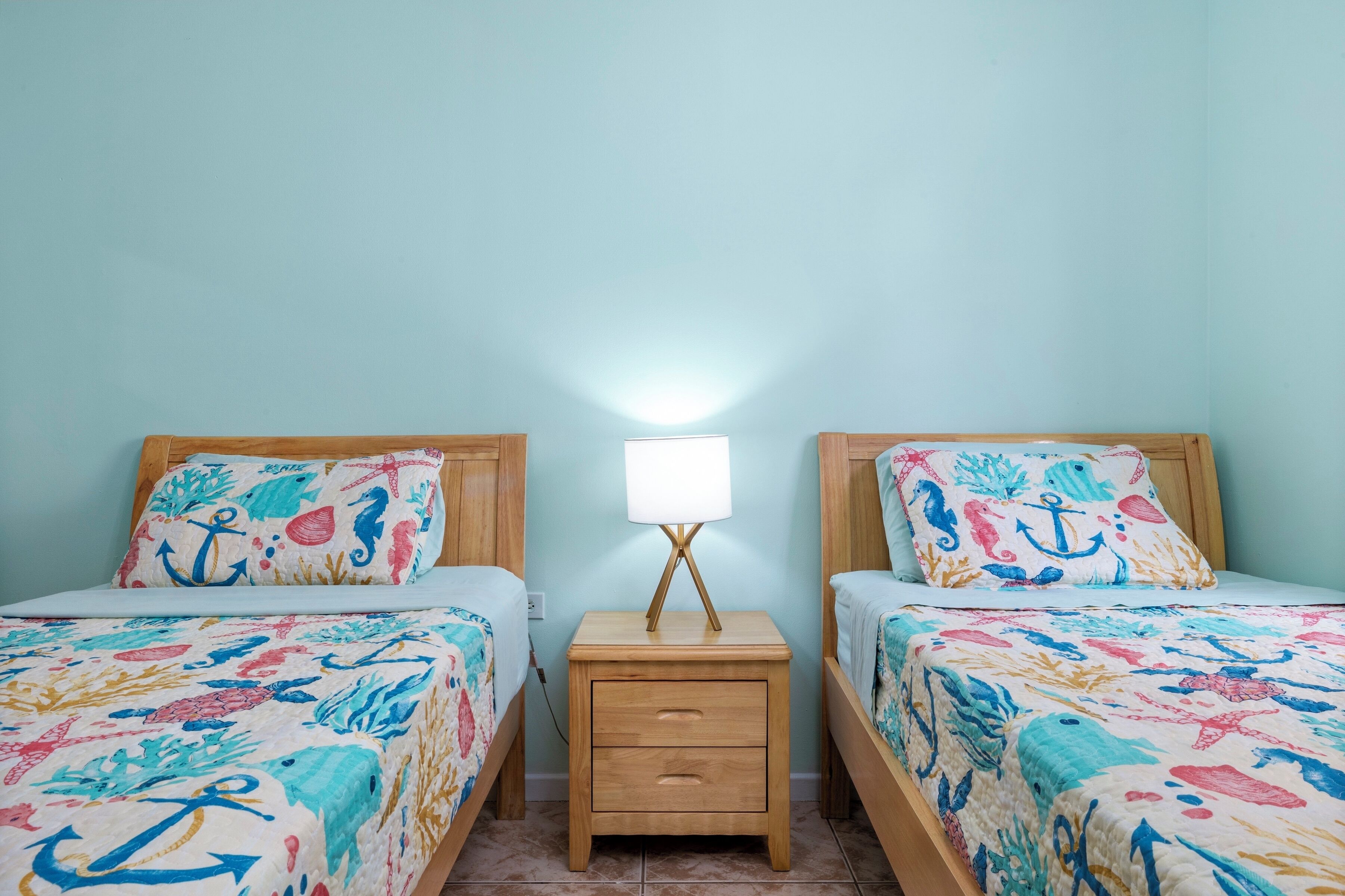 A twin room with two beds, each with a nautical-themed bedspread, a bedside table with a lamp, and a nightstand with drawers.