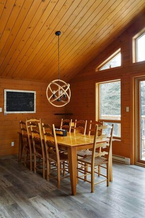 Un coin repas en bois avec une table ronde et des chaises, un luminaire suspendu et de grandes fenêtres.
