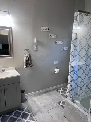 Ein Badezimmer mit grauer Wand, einem Spiegel, einem Waschbecken, einem Handtuch, einem Toilettenpapierhalter und einer Duschvorhang.