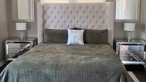 Ein Schlafzimmer mit einer gepolsterten Kopfteile, einem Bett mit einer strukturierten, grünen Tagesdecke, zwei Nachttischen mit Lampen und einem Deckenventilator.