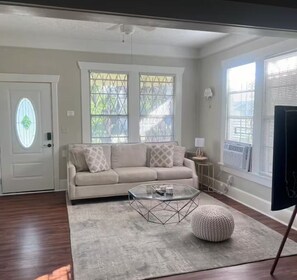 Ein Wohnzimmer mit einem beigen Sofa, einem Glastisch, einem runden Ottomanen und einer Tür mit einem Buntglasfenster.