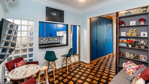 Een kamer met een blauwe wand, een plafondventilator, een zwart-wit betegelde spatwand, een ronde houten tafel met twee stoelen, een boekenkast en een vloer met patroon.
