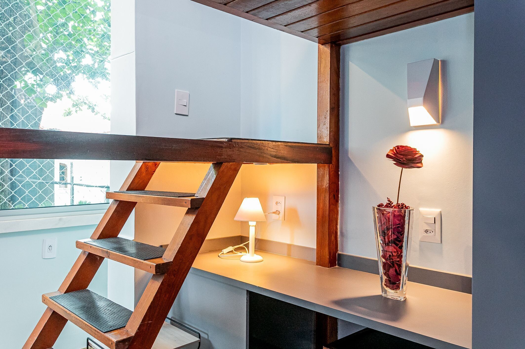 A staircase with a wooden handrail, a white wall-mounted lamp, and a vase with a single red rose.