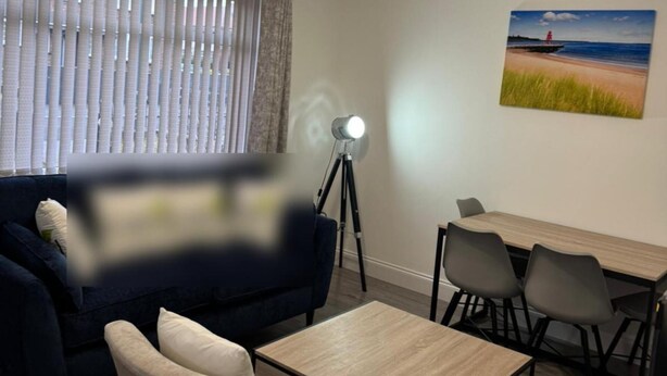 Una sala de estar con un sofá, una mesa de centro y una mesa de comedor con sillas. Hay una lámpara sobre un atril.