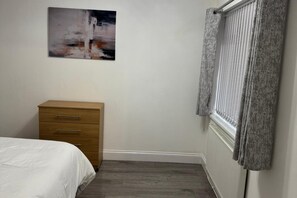 A bedroom with a wooden dresser, a bed, a window with grey curtains, and a painting on the wall. - Bungalow - Sleeps 2 - Parking - Garden (Jarrow)