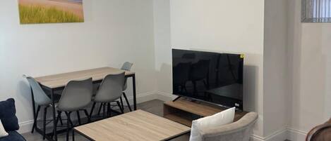 A modern living room with a wooden dining table, grey chairs, a flat-screen TV, and a sofa.