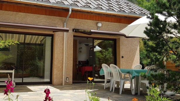 A house with a tiled roof, a patio with a table and chairs, and a garden with various plants and flowers. - Au fil du Réguinier – Seasonal Rental Cottage (Briançon)