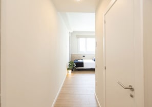 Un pasillo estrecho con una cama al final, una planta y una puerta con picaporte.