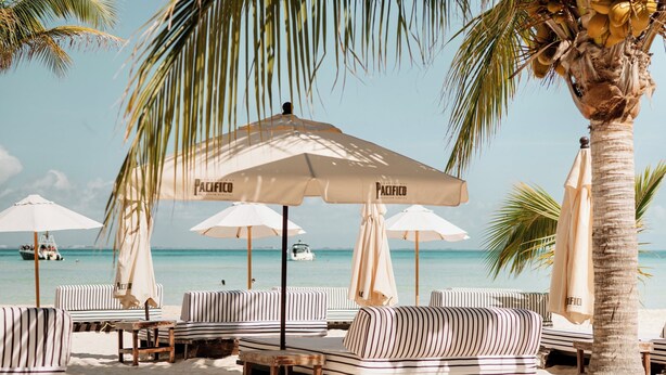 A beachside lounge area with striped lounge chairs and umbrellas, a palm tree, and a clear view of the ocean. - Wake up to turquoise waters at Isla Bliss 🌴 — a luxury 3-bedroom oceanfront condo just minutes from Isla Mujeres’ best dining and beaches. Enjoy a 90" Smart TV, stunning views, fast WiFi, and easy self-check-in for the perfect island escape. 🌊✨ (Isla Mujeres)