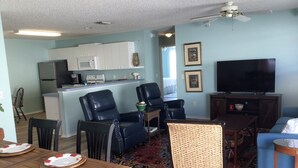 A living room with a TV, two leather chairs, a dining table with plates, a kitchen area with cabinets, and a ceiling fan. - Live the Villages lifestyle in  this 2 bedroom beauty (The Villages)