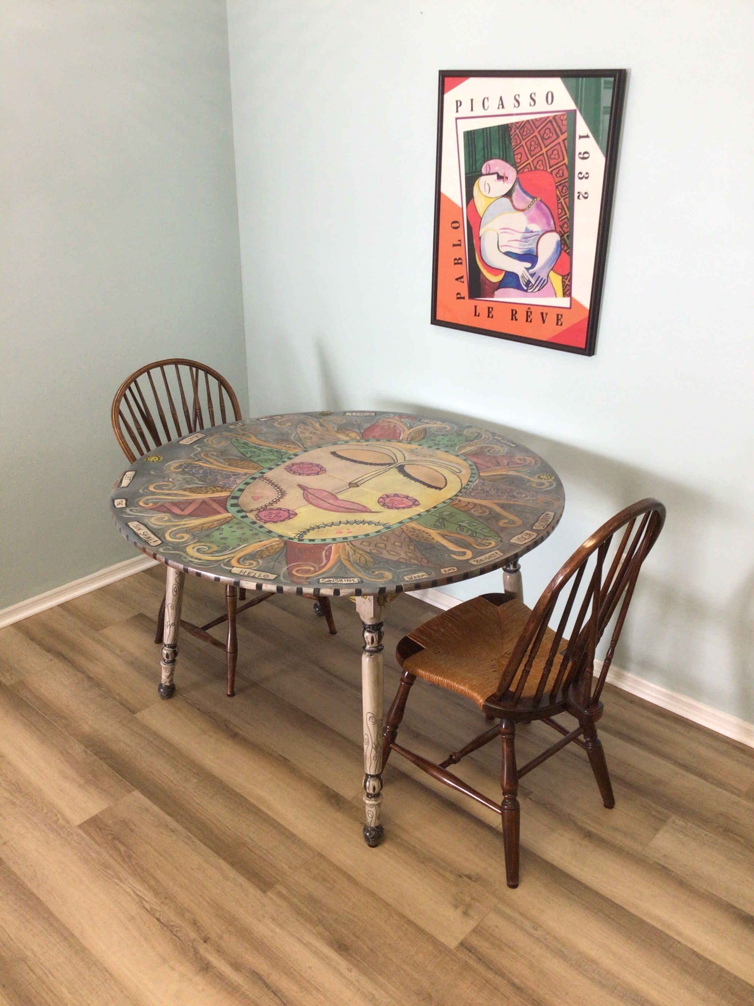 A round dining table with a colorful mosaic top, surrounded by four wooden chairs. A framed artwork of a rooster is mounted on the wall above the table.