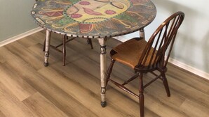 A round dining table with a colorful mosaic top, surrounded by four wooden chairs. A framed artwork of a rooster is mounted on the wall above the table. - Live the Villages lifestyle in  this 2 bedroom beauty (The Villages)