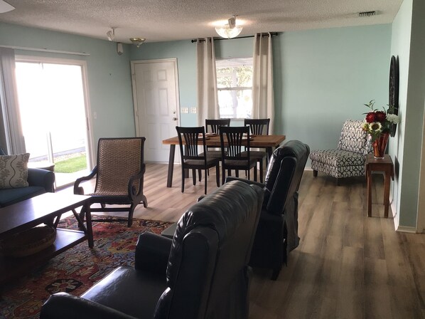 A living room with a sofa, a leather chair, a dining table with chairs, a patterned rug, and a vase with flowers on a side table.