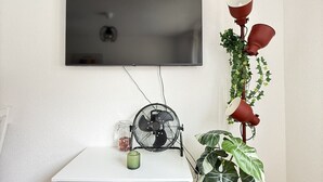 Ett rum med en platt-TV som är monterad på väggen, en vit byrå, en liten fläkt, en röd golvlampa och en krukväxt.