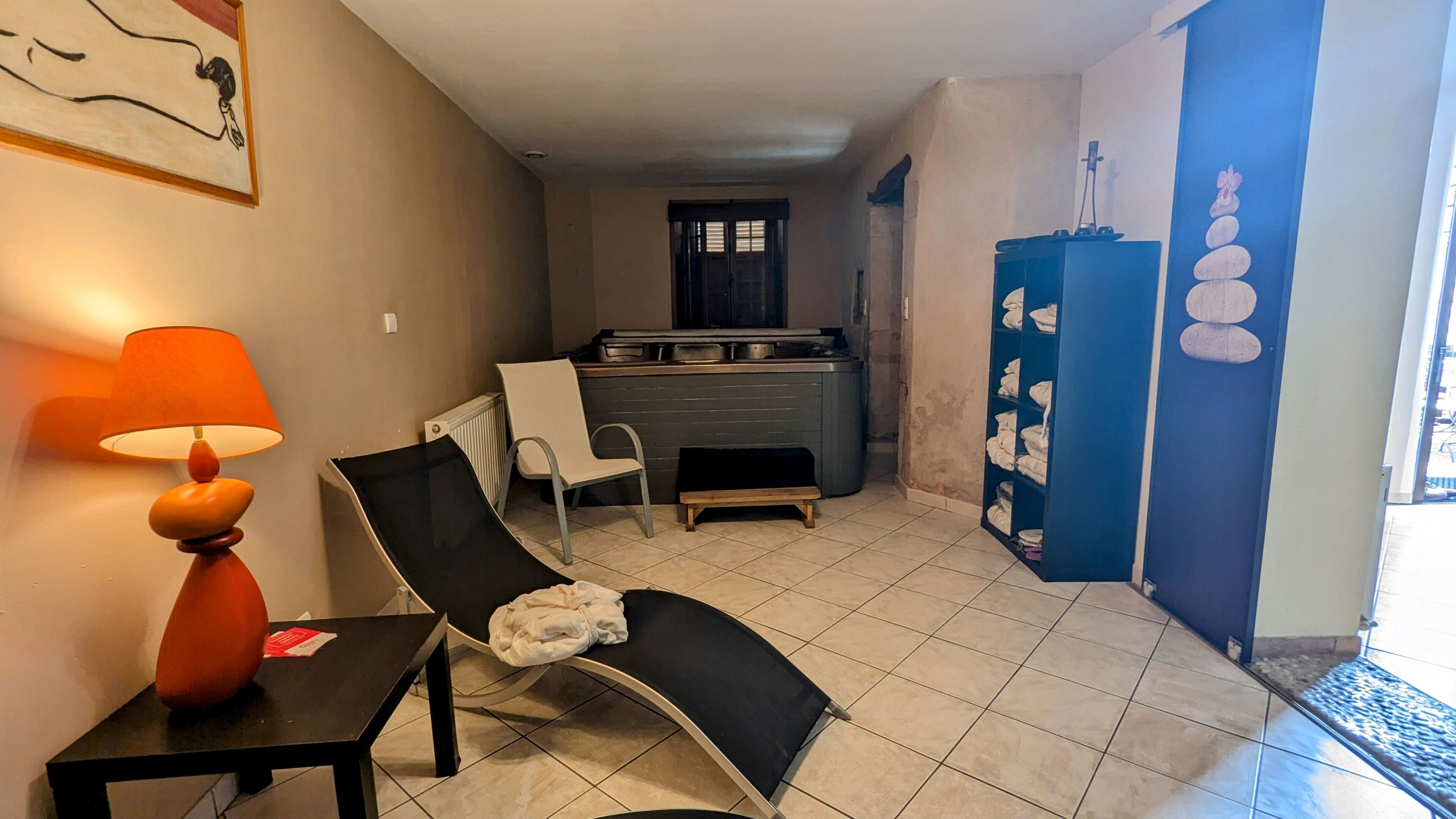 A living room with a black lounge chair, a table with a lamp, a radiator, a kitchen area with a stove, and a blue wall with stacked towels.