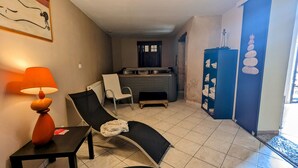 Una sala de estar con un sillón reclinable negro, una mesa con lámpara, un radiador, una zona de cocina con estufa y una pared azul con toallas apiladas.
