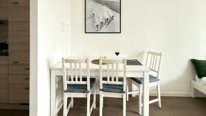 Un coin repas avec une table et des chaises blanches, un tableau encadré au mur et une petite table sur laquelle se trouve un verre de vin rouge.