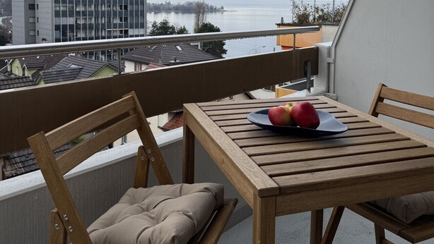 Un balcon avec une table et des chaises en bois, un coussin et une assiette de fruits. On aperçoit un grand immeuble et une étendue d’eau en arrière-plan.