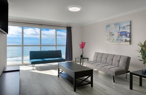 Ein modernes Wohnzimmer mit großem Fenster mit Blick auf den Ozean, ein blaues Sofa, ein graues Sofa, ein Couchtisch und ein Gemälde von einem Lieferwagen und einem Surfbrett.