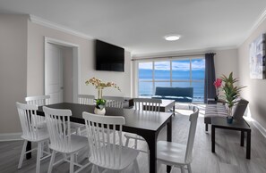 Ein Essbereich mit großem Tisch und weißen Stühlen, einem Flachbildfernseher an der Wand und Blick auf das Meer durch ein großes Fenster.