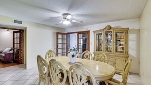 Uma sala de jantar com uma mesa redonda, seis cadeiras, um ventilador de teto e um armário de madeira com portas de vidro.