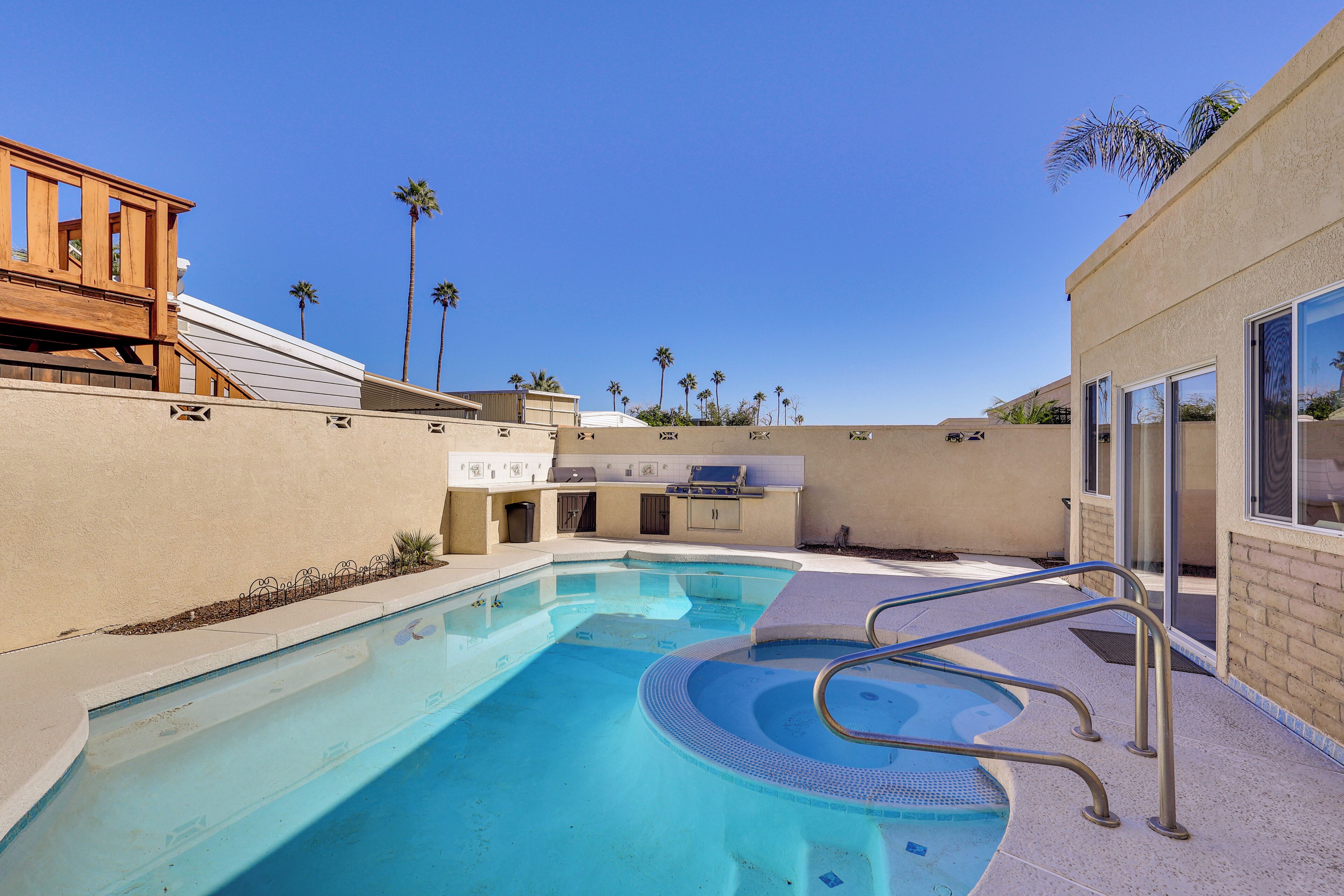 A swimming pool with a wooden deck and metal railings, surrounded by palm trees and a clear blue sky.