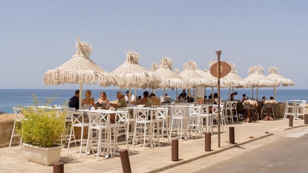 Een terras met overdekte zitplaatsen onder rieten parasols, witte stoelen en uitzicht op zee.
