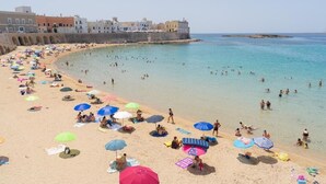 Een strand met mensen die zwemmen en zonnebaden, parasols en gebouwen op de achtergrond.