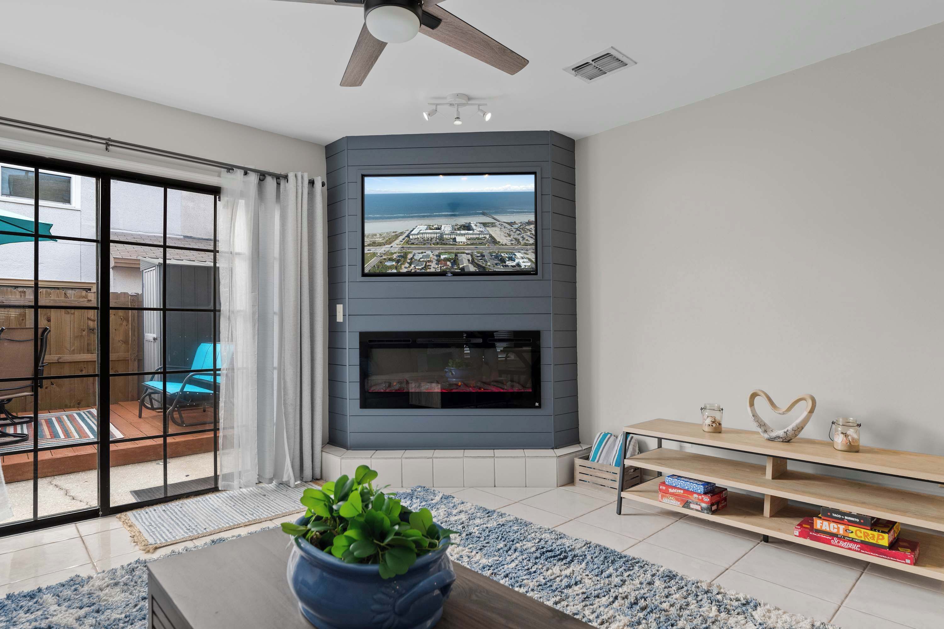 Un salon moderne avec un grand téléviseur fixé sur un mur gris, une cheminée en dessous et une porte-fenêtre coulissante donnant sur une terrasse où se trouve une chaise bleue.