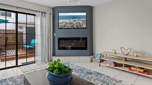 Ein modernes Wohnzimmer mit einem großen Fernseher an einer grauen Wand, einem Kamin darunter und einer Schiebetür zu einer Terrasse mit einem blauen Stuhl.