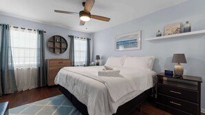 Ein Schlafzimmer mit einem Bett, einem Nachttisch, einem an der Wand befestigten Regal, einem Deckenventilator und einem Fenster mit Vorhängen.