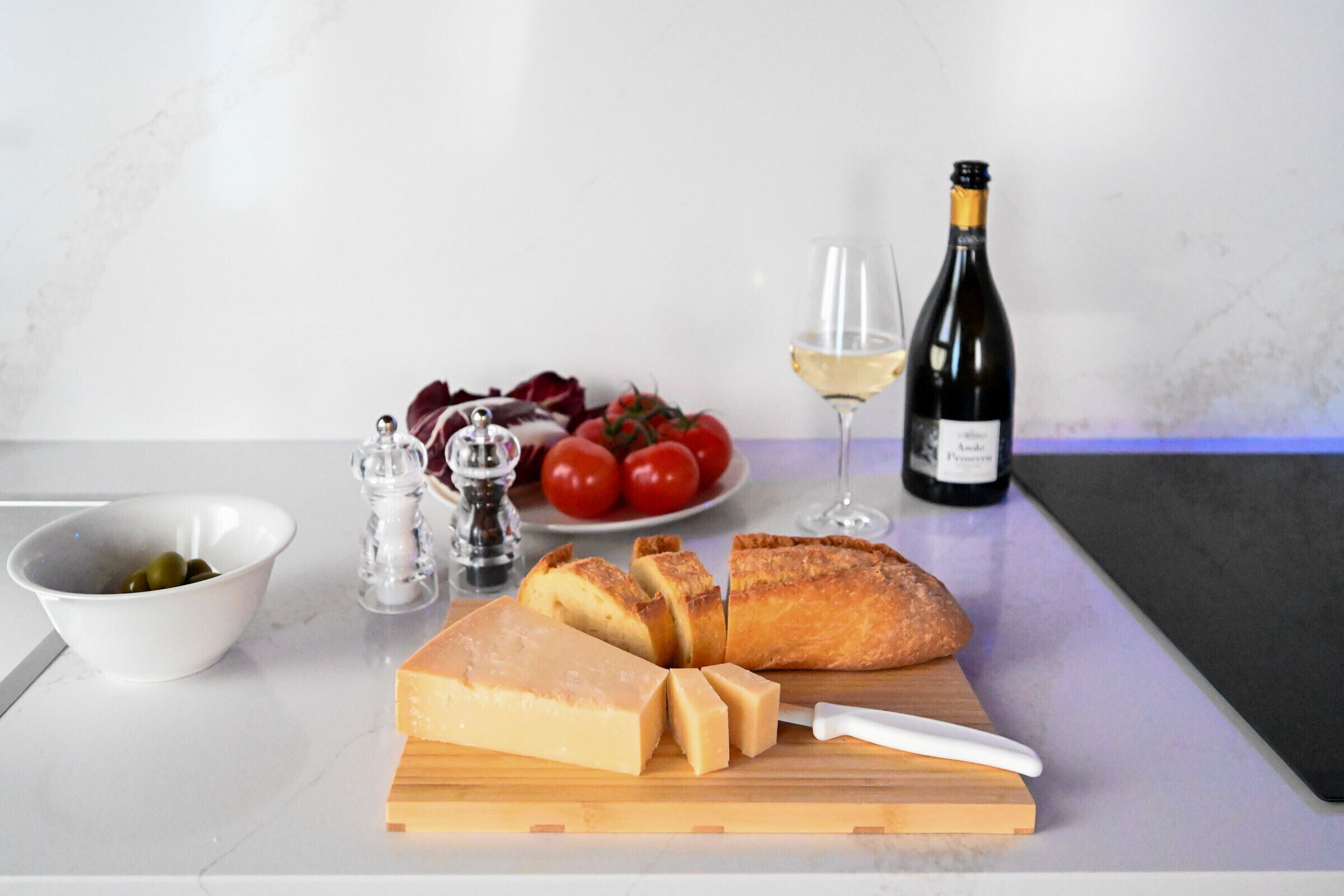 Un piano di lavoro da cucina con una pagnotta, una bottiglia di vino, un piatto di pomodori, una ciotola di olive e un coltello su un tagliere.