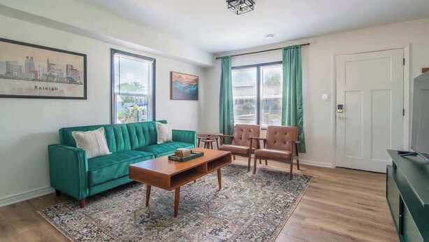 Ein Wohnzimmer mit einem grünen Sofa, einem hölzernen Couchtisch und zwei Stühlen. Auf dem Boden liegt ein gemustertes Teppich, an der Wand hängt ein gerahmtes Stadtbild und rechts ist eine Tür zu sehen.