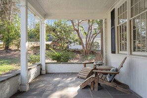 Une terrasse couverte avec deux fauteuils Adirondack en bois, une petite table et une vue sur un jardin et les maisons voisines.