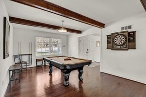Une table de billard dans une pièce au plancher en bois, un jeu de fléchettes et une fenêtre avec des stores.
