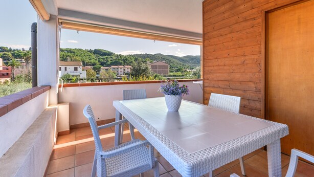 Un balcone con un tavolino e sedie in vimini bianco, una pianta in vaso e vista su un paesaggio verde con edifici.