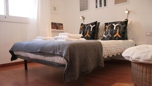 Uma cama bem arrumada com um cobertor cinza e duas almofadas com padrões geométricos, uma cadeira de vime e uma pequena mesa com um abajur.