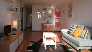 Uma sala de estar moderna com um sofá cinzento, uma escrivaninha de madeira, uma mesa de centro branca e uma área de jantar com cadeiras laranja.