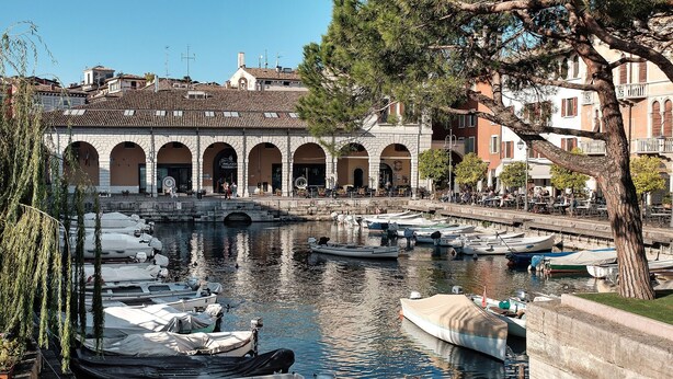 A canal with boats docked, a stone building with arches, and a tree in the foreground. - Apartment 'Il Vagabondo' with Wi-Fi and Air Conditioning (Volta Mantovana)