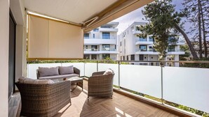 Een balkon met rieten meubilair, een glazen tafel en uitzicht op moderne gebouwen en bomen.