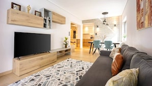 Een moderne woonkamer met een flatscreen televisie, houten planken, een kamerkleed met dessin en een grijze bank met decoratieve kussens.