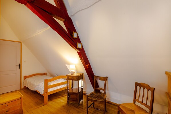 Um quarto pequeno e acolhedor com teto inclinado, uma cama de solteiro, uma cadeira de madeira, uma mesa pequena com luminária e uma porta.