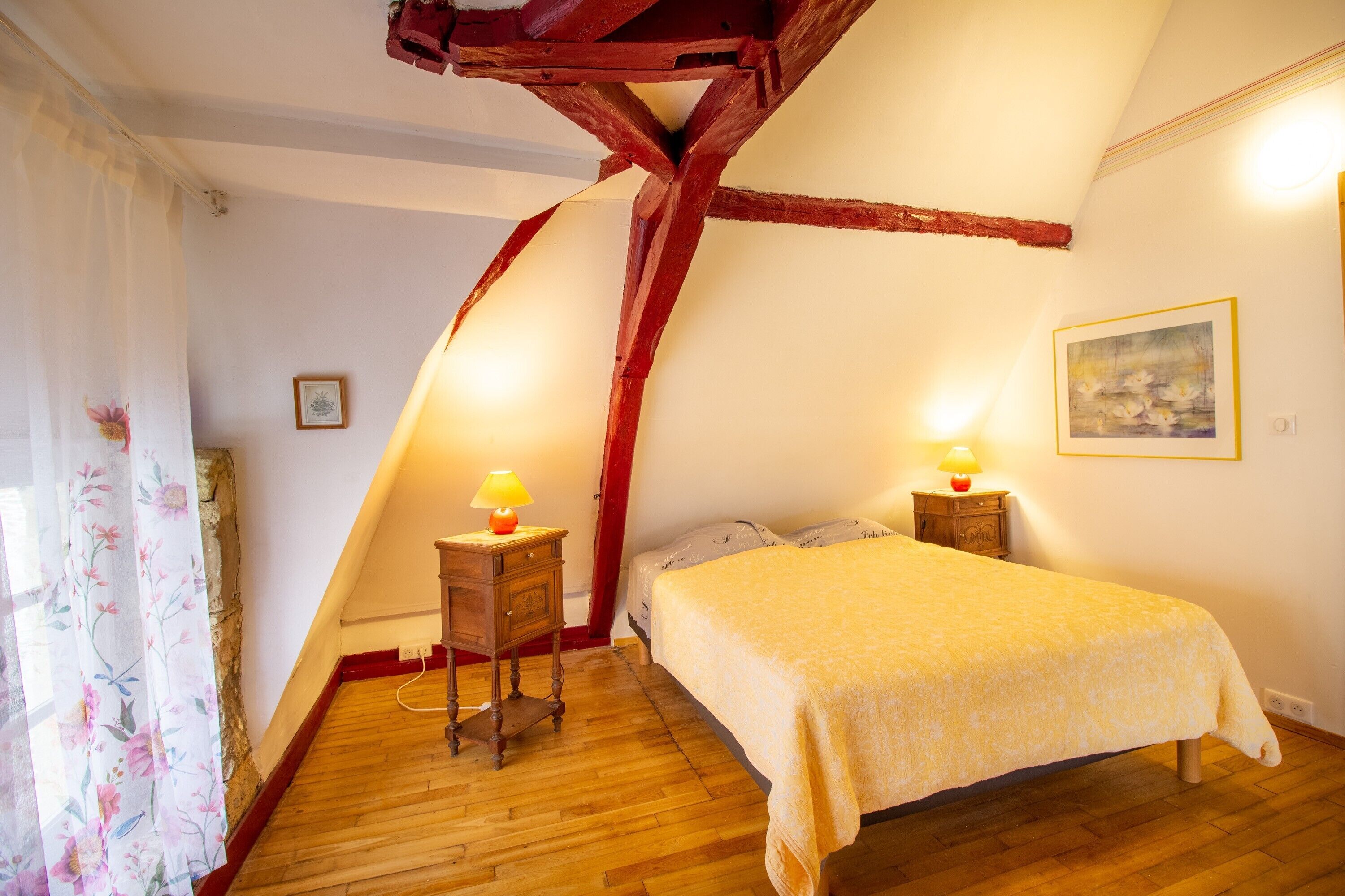 Une chambre avec un plancher en bois, un lit recouvert d’une couvre-lit jaune, deux lampes de chevet, une table de chevet en bois, une poutre rouge, un tableau au mur et un rideau à motifs floraux.