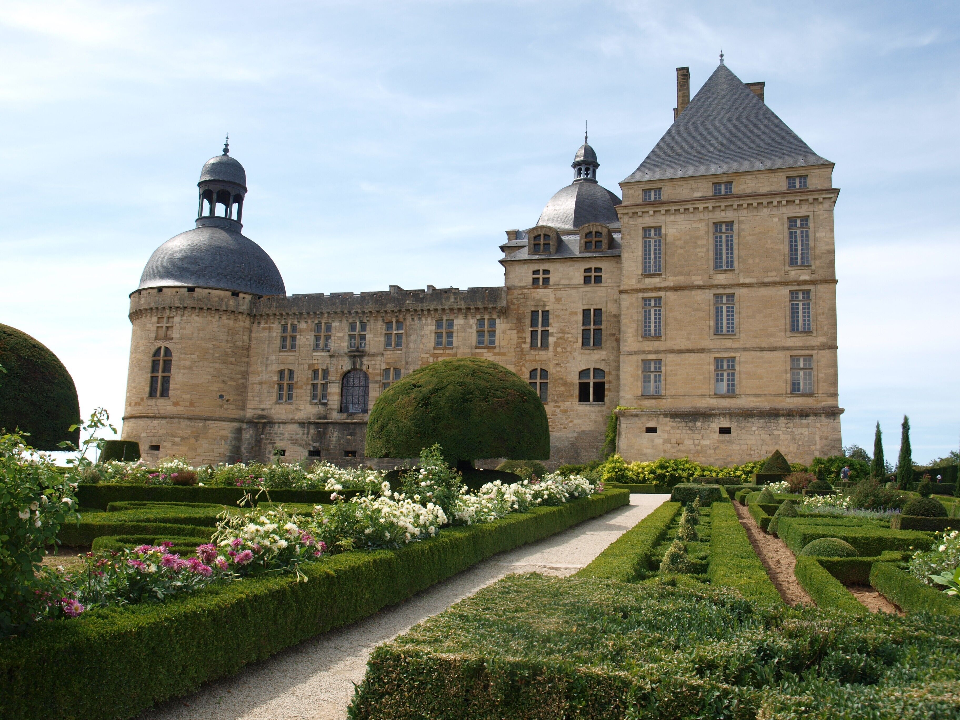 A historic building with a central dome, a large tower, and a formal garden with hedges and flower beds.