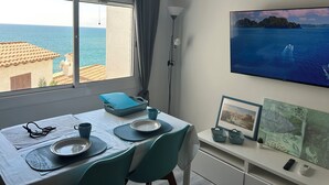 A room with a dining table set for two, a TV on the wall, and a view of the sea through the window. - APARTMENT WITH FANTASTIC SEA VIEW NEXT TO THE BEACH SWIMMING POOL PARKING WIFI (SITGES)