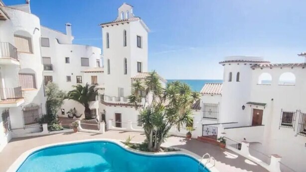 A swimming pool surrounded by white buildings with balconies, a clear sky, and a view of the sea. - APARTMENT WITH FANTASTIC SEA VIEW NEXT TO THE BEACH SWIMMING POOL PARKING WIFI (SITGES)