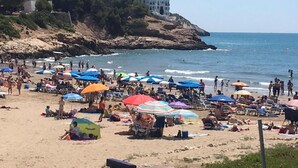 A beach with people sunbathing, umbrellas, and a rocky cliff with buildings. - APARTMENT WITH FANTASTIC SEA VIEW NEXT TO THE BEACH SWIMMING POOL PARKING WIFI (SITGES)