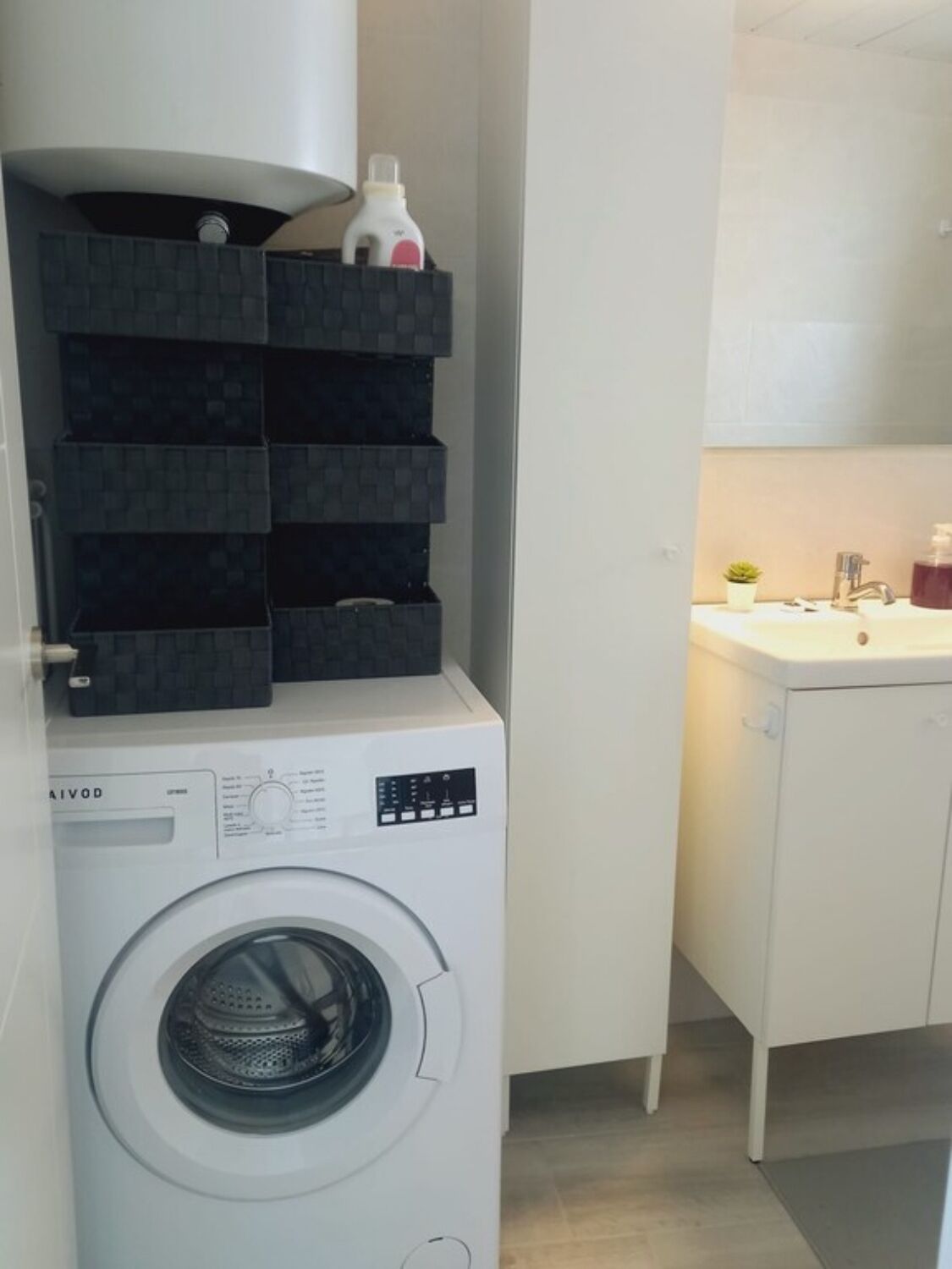 Ein kleiner Waschraum mit einer Waschmaschine mit Frontlader, einem Regal mit Aufbewahrungsbehältern und einem kleinen Waschbecken mit Topfpflanze.