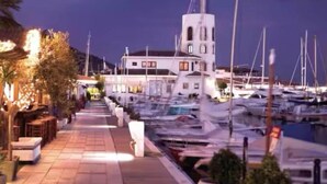 A marina with boats docked, a white building with a tower, and a paved walkway with tables and chairs. - APARTMENT WITH FANTASTIC SEA VIEW NEXT TO THE BEACH SWIMMING POOL PARKING WIFI (SITGES)
